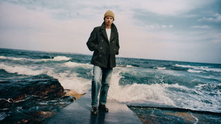 Man wearing the Noah x Gloverall peacoat open over jeans, standing on a rocky shoreline with waves breaking behind him