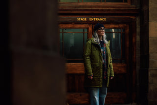 A woman with gray hair stands in front of a dark wooden stage entrance door, wearing an olive green parka, a dark beanie, and blue jeans.