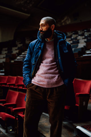 A man with a beard wearing a blue puffer jacket over a pink knit sweater and brown corduroy pants, standing in a dimly lit auditorium with red seats.