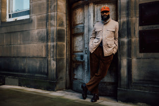 A man wearing a beige jacket, brown trousers, and an orange cap leans against a weathered wooden door in a stone building.