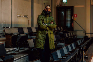 A man wearing a long olive green hooded puffer coat stands with arms crossed in an empty indoor auditorium or lecture hall with rows of dark chairs.