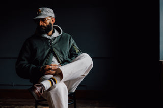 A man wearing a dark green bomber jacket, white sweatpants, a gray hoodie, and a gray cap with a yellow logo sits on a stool in a dimly lit studio setting.