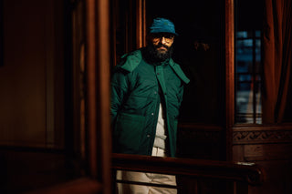 A man wearing a dark green puffer jacket, a blue cap, and glasses stands in a dimly lit, traditional wooden interior.